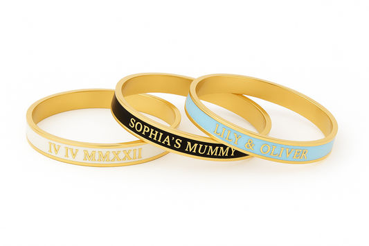3 enamel bangles with different customised text on a white background. White and gold bangle reads 'IV IV MXXIII', black and gold bangle reads 'Sophia's Mummy', pale blue and gold bangle reads 'Lily & Oliver'